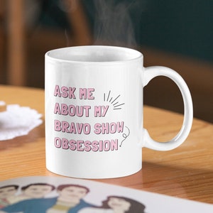 May include: White ceramic coffee mug with a pink graphic that says "Ask me about my Bravo show obsession".