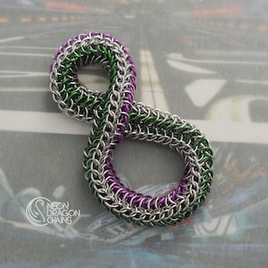 May include: A chainmail infinity symbol pendant crafted from interlocking silver, vibrant green, and deep purple metal rings. The design features a continuous loop pattern with a smooth, woven texture. The text NEON DRAGON CHAINS is visible.