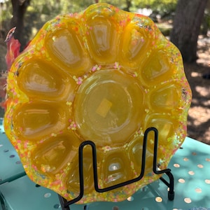 May include: Yellow resin egg dish with a sparkly finish. The dish has 10 individual compartments for holding eggs. The dish is on a black metal stand.