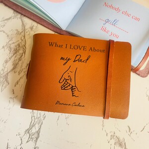 May include: A tan leather journal with the text "What I LOVE About my Dad" and a drawing of two hands holding each other. The journal has a leather strap closure. An open book with text is visible in the background.