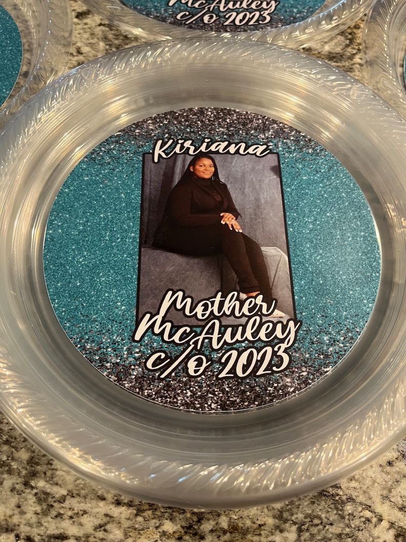 May include: A clear plastic plate featuring a central circular insert with a teal glitter background. The insert displays a photograph of a person sitting, with the text Kiriana above and Mother McAuley c/o 2023 printed in white script below.