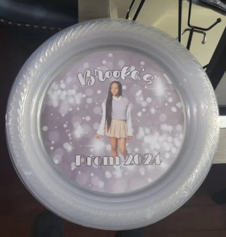 May include: A round, clear plastic plate featuring a central circular insert with a photograph of a person wearing a light-coloured sweater and skirt. The text "Brooke's" appears at the top and "Prom 2024" at the bottom against a bokeh background.