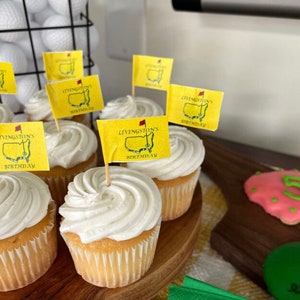 May include: A close-up of cupcakes decorated with white frosting and topped with yellow flags that say "Livingston's Birthday". The flags are attached to toothpicks and are placed in the frosting. The cupcakes are on a wooden surface.