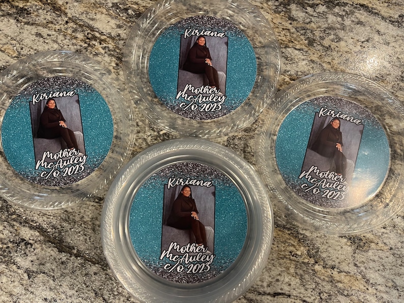 May include: Four circular clear plastic containers featuring a central insert with a blue and silver glitter background. Each insert displays a photograph of a person and the text Kiriana Mother McAuley c/o 2023 printed in a black script font.