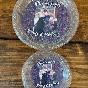 May include: Two clear plastic circular plates featuring a central purple glitter insert. The insert displays a photograph of two individuals in formal attire with the text Prom 2023 and Kacy & Kelsey printed in white script lettering on the surface.