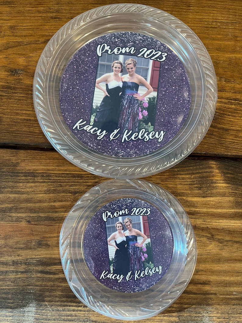 May include: Two clear plastic circular plates featuring a central purple glitter insert. The insert displays a photograph of two individuals in formal attire with the text Prom 2023 and Kacy & Kelsey printed in white script lettering on the surface.