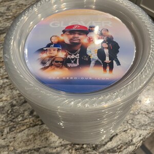 May include: A stack of clear plastic plates featuring a circular graphic insert on the top plate. The graphic displays a collage of individuals, the text "JUSTEN GLOVER" at the top, and the text "OUR HERO OUR ICON" at the bottom.