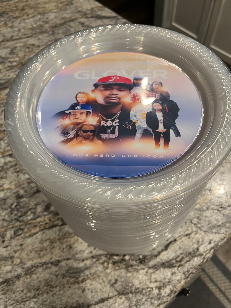May include: A stack of clear plastic plates featuring a circular graphic insert on the top plate. The graphic displays a collage of individuals, the text "JUSTEN GLOVER" at the top, and the text "OUR HERO OUR ICON" at the bottom.