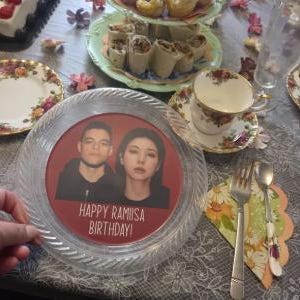 May include: A clear, circular plastic plate featuring a red centre insert with a photograph of two people and the text "HAPPY RAMIISA BIRTHDAY!" printed in white. The plate has a textured, ridged border and is set on a table with floral china.