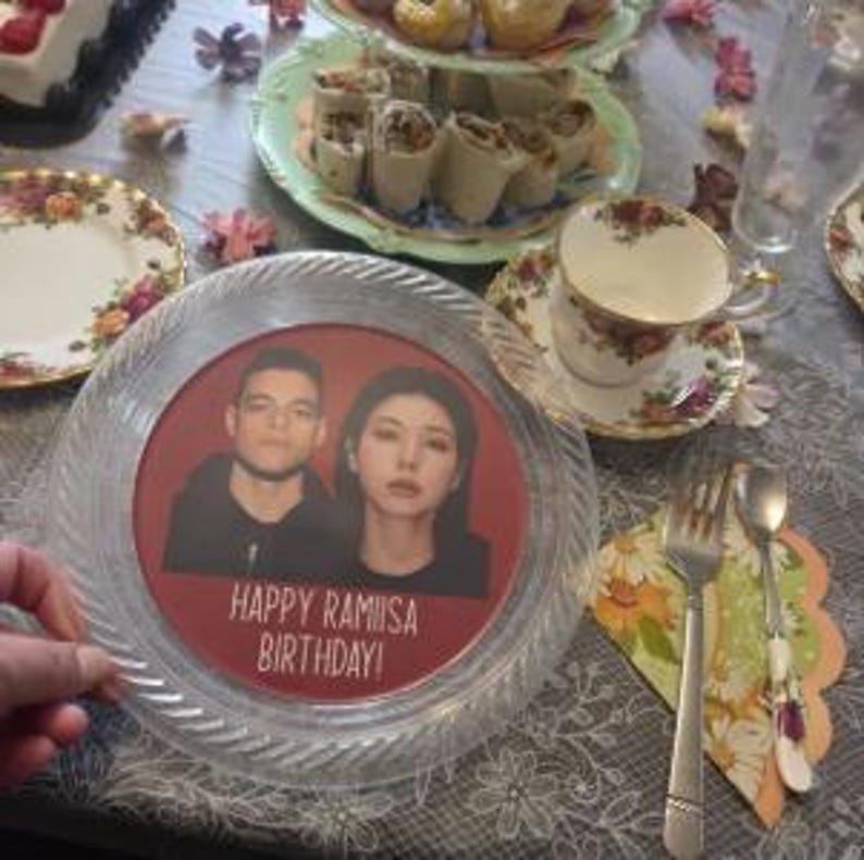 May include: A clear, circular plastic plate featuring a red centre insert with a photograph of two people and the text "HAPPY RAMIISA BIRTHDAY!" printed in white. The plate has a textured, ridged border and is set on a table with floral china.
