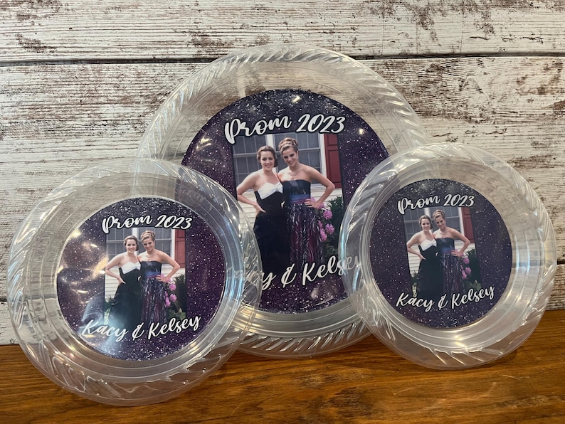 May include: Three clear plastic plates of varying sizes, each featuring a circular insert with a purple glitter background. The inserts display a photograph of two people, the text Prom 2023, and the names Kacy & Kelsey printed in white script.