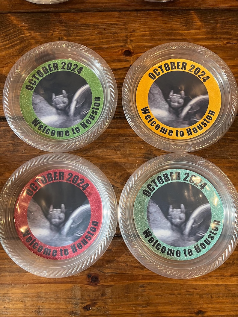 May include: Four round clear plastic containers featuring circular labels with a black and white ultrasound image in the centre. The labels have glittery borders in green, yellow, red, and teal, with the text OCTOBER 2024 Welcome to Houston.