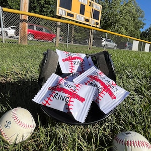 May include: A black baseball cap with five small white bags inside. The bags have red stitching and the text "CHAMPIONSHIP RING" printed on them. Two white baseballs are in the grass below the cap.