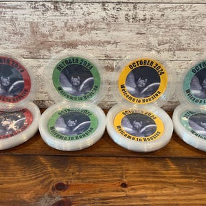 May include: Four stacks of clear plastic plates featuring circular inserts with a bat graphic and the text OCTOBER 2024 Welcome to Houston. The inserts come in red, green, yellow, and teal glitter borders, arranged in two rows on a wooden surface.