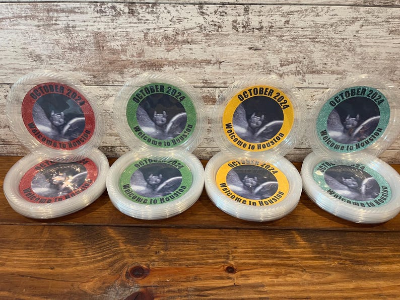 May include: Four stacks of clear plastic plates featuring circular inserts with a bat graphic and the text OCTOBER 2024 Welcome to Houston. The inserts come in red, green, yellow, and teal glitter borders, arranged in two rows on a wooden surface.