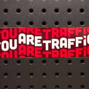 Puede incluir: Una pegatina roja y blanca con el texto "You are traffic" repetido tres veces.