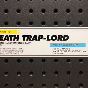 Puede incluir: Una etiqueta blanca y amarilla con el texto "DEATH TRAP-LORD" y otro texto, incluyendo "TAKE ONE INJECTION ONCE DAILY (420MG)". La etiqueta está sobre una superficie gris oscuro con agujeros espaciados.