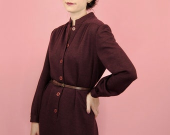 May include: A burgundy dress with long sleeves and a button-up front. The dress is cinched at the waist with a brown belt. The dress has a mandarin collar and falls to the knees. The background is a solid pink.