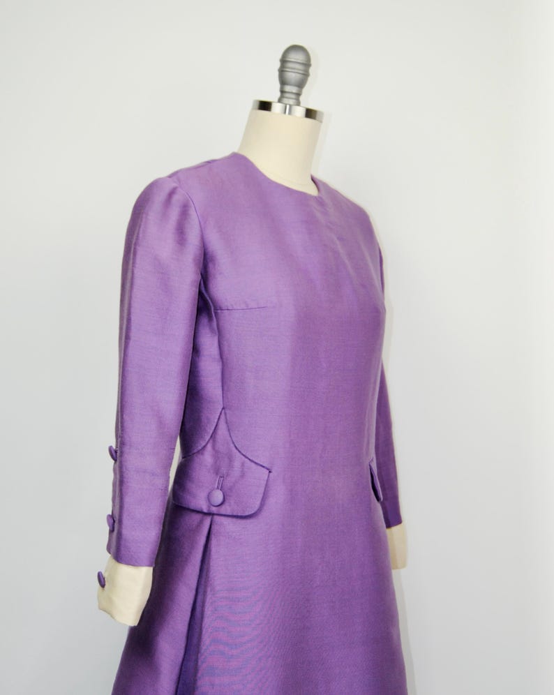May include: A long-sleeved, purple dress with a round neckline and button details on the cuffs and pockets.