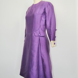 May include: A purple vintage dress with long sleeves and a button detail on the side. The dress has a fitted bodice and a slightly flared skirt.