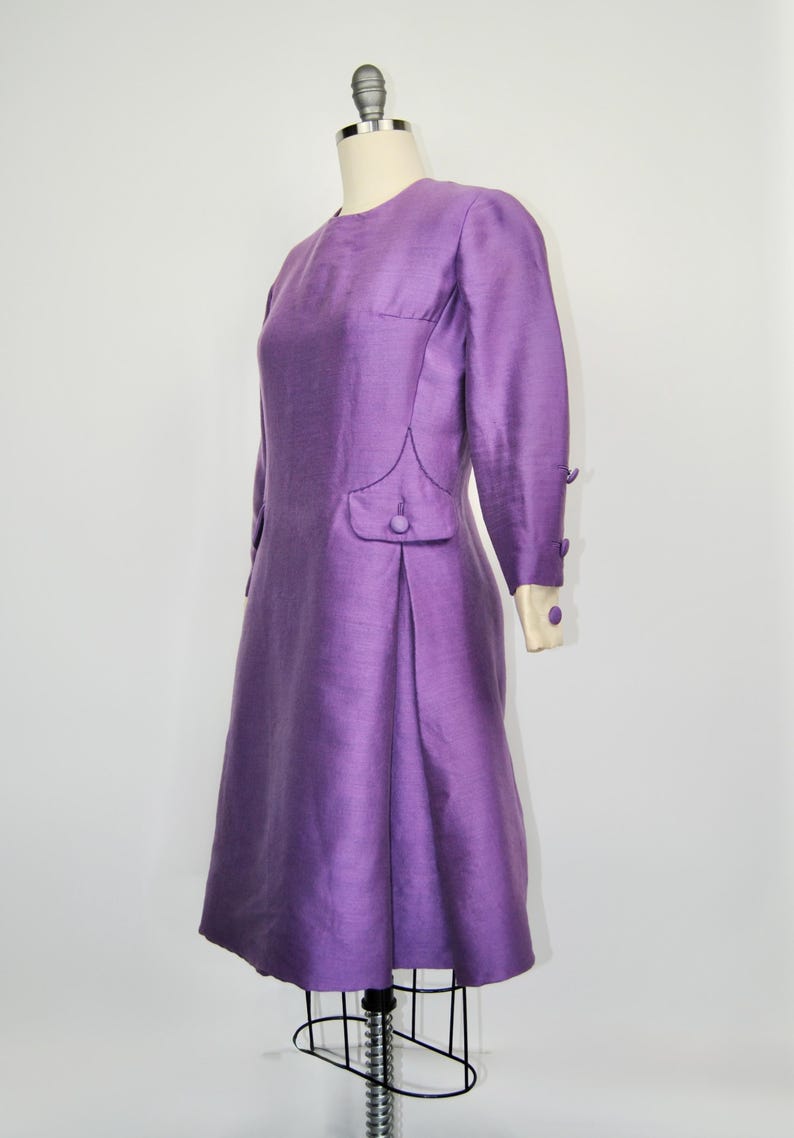May include: A purple vintage dress with long sleeves and a button detail on the side. The dress has a fitted bodice and a slightly flared skirt.