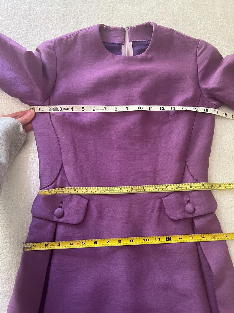 May include: A purple dress with long sleeves and a round neckline. The dress has two pockets on the front and a zipper in the back. The dress is measured with a tape measure, showing the length of the dress is 36 inches.