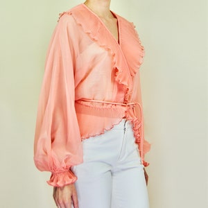 May include: A peach-colored sheer blouse with a ruffled collar and long sleeves. The blouse has a wrap-style front and a tie at the waist.