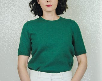 May include: A woman wearing a green short-sleeved sweater and white pants. She is standing in front of a white background.