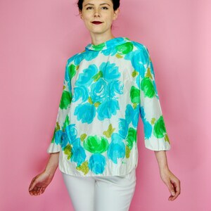 May include: A woman wearing a white blouse with a blue and green floral print and white pants. She is standing in front of a pink background.