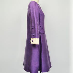 May include: A purple dress with long sleeves and white cuffs. The dress has a fitted bodice and a flared skirt. The dress has buttons on the sleeves and a seam down the side.