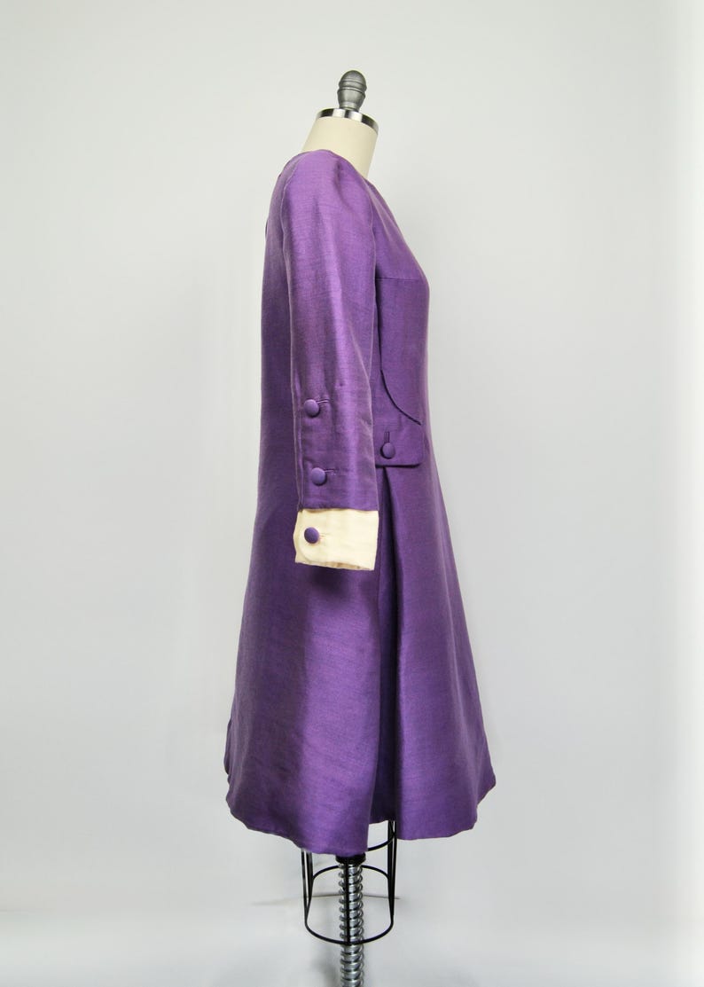 May include: A purple dress with long sleeves and white cuffs. The dress has a fitted bodice and a flared skirt. The dress has buttons on the sleeves and a seam down the side.