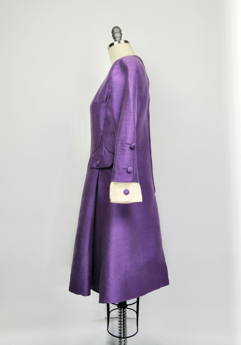 May include: A purple dress with a white trim on the sleeves and a button detail. The dress is on a mannequin.