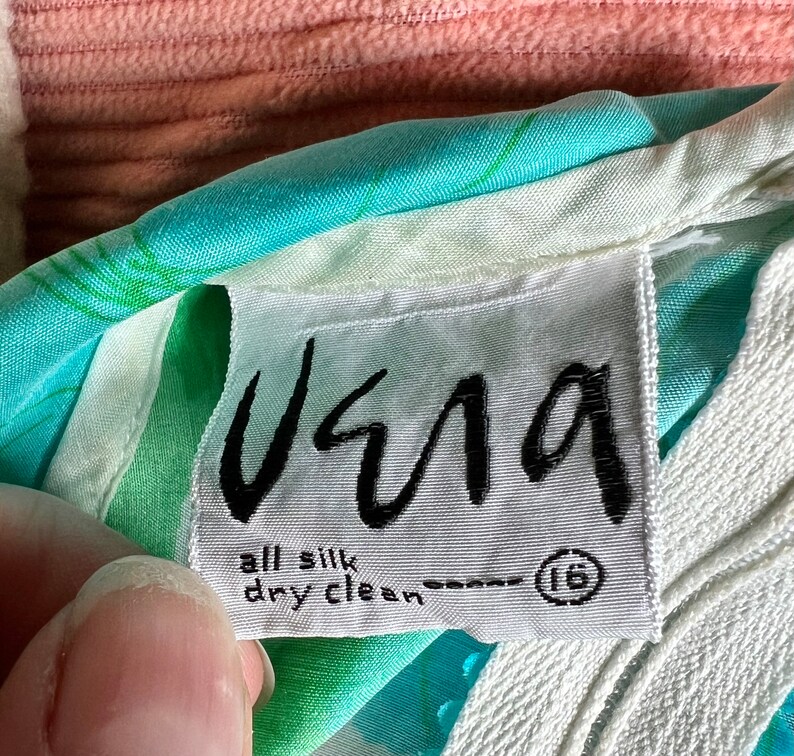 May include: A white tag with black text that reads "Usma" and "all silk dry clean" with a size 16 in a circle. The tag is attached to a turquoise and white patterned fabric.