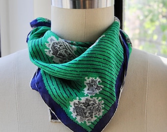May include: A green and blue silk scarf with a white and black pattern of buildings. The scarf has a striped pattern and a navy blue border.