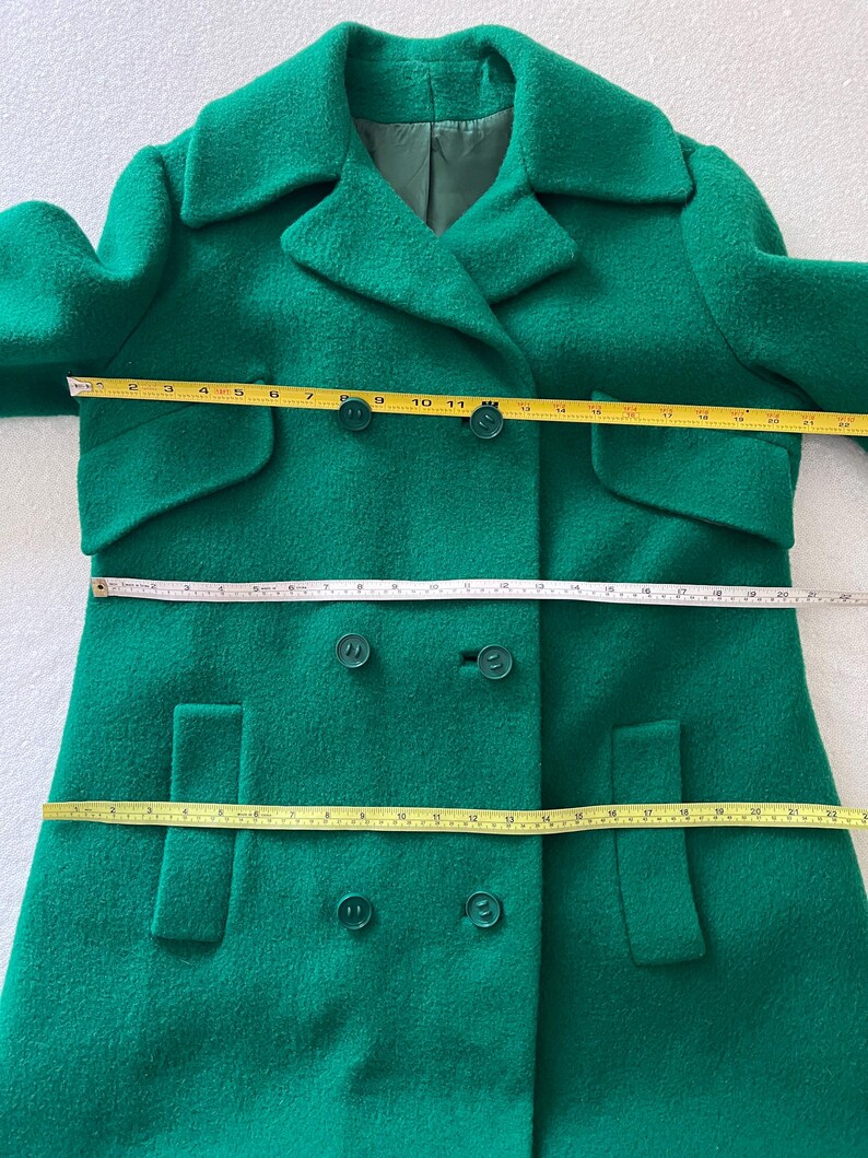 Pu&ograve; includere: Un cappotto di lana verde a doppio petto con due tasche su ogni lato e quattro bottoni sul davanti.