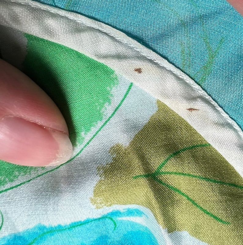 May include: Close-up of a white fabric with a green, blue, and brown abstract pattern. The fabric has a white trim with two small brown stains.