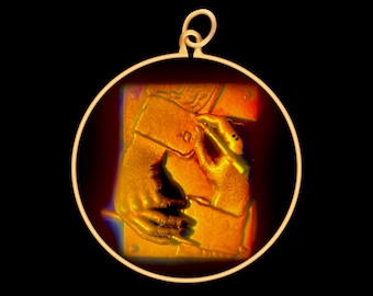 Può includere: Un ciondolo d'oro con una cornice circolare che presenta un'immagine dettagliata di una mano che tiene una penna. L'immagine è in una tavolozza di colori caldi, arancio e rosso.