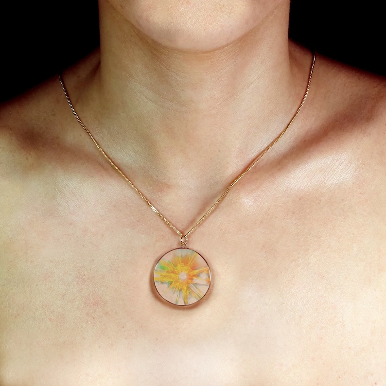K&ouml;nnte beinhalten: Eine goldene Kettenkette mit einem runden Anh&auml;nger mit einem abstrakten Design in Gelb-, Orange- und Gr&uuml;nt&ouml;nen.