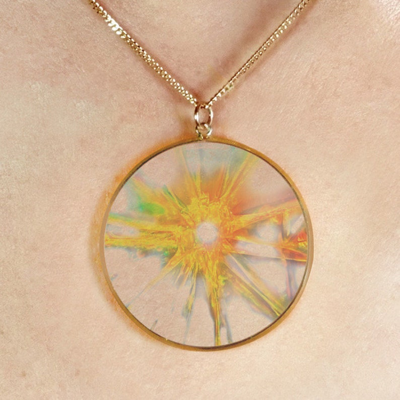 May include: A gold chain necklace with a round pendant featuring a colorful abstract design with a yellow center.