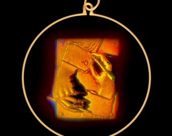 Può includere: Un ciondolo d'oro con una cornice circolare e un'immagine dettagliata di una mano che tiene una penna. L'immagine è in uno schema di colori caldi, arancione e giallo.