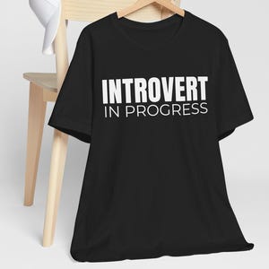 May include: A black short-sleeved t-shirt featuring the white text INTROVERT IN PROGRESS printed on the chest. The shirt is displayed on a wooden hanger, and a plain white baseball cap is draped over the top corner of the wooden chair back.