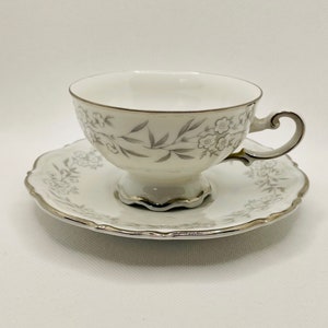 May include: A white teacup and saucer set with a silver floral design. The cup has a scalloped edge and a handle. The saucer has a scalloped edge.