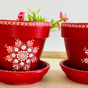 May include: Two red terracotta plant pots with matching saucers, each featuring a hand-painted white and light pink mandala-style floral pattern on the front and a dotted border along the top rim and the edge of the saucer base.