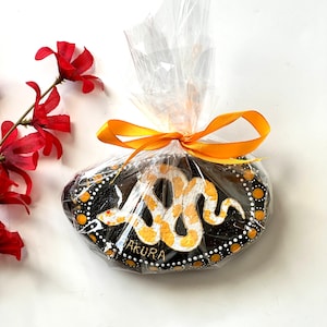 May include: A hand-painted black stone with a gold and white snake design, wrapped in clear cellophane and tied with a gold ribbon. The word "Sakura" is written on the stone. Red flowers are in the background.