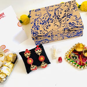 May include: A gift set featuring a rectangular box with a blue floral pattern, a white card with red text, a pair of gold-tone earrings with red and yellow accents on a black cushion, two small vials, a chocolate bar, and a decorative tray with two bowls.
