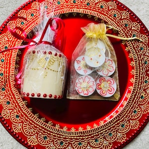 May include: A red decorative plate with gold filigree holds a wrapped candle and a small bag of floral-designed tea lights. The candle is cream-colored with gold script and red accents. The tea lights have red and gold floral designs.