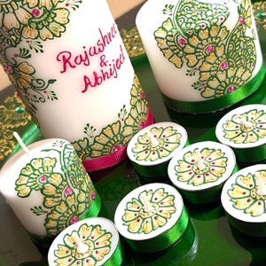 Puede incluir: Velas blancas con intrincados diseños florales verdes y dorados, acentuados con detalles rosas. La vela más grande tiene los nombres "Rajashree & Abhijeet" en rosa. También hay velas de té con diseños a juego y cinta verde.