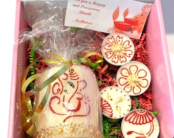 May include: A pink gift box containing a large pillar candle wrapped in clear plastic with a gold ribbon, featuring a red Ganesha design and rhinestones. Four small round tea light candles with red and gold patterns are included with a card.