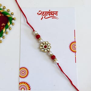 May include: A Rakhi bracelet featuring a central flower-shaped charm with white stones and a red center, flanked by red and white beads on a red thread. The bracelet is displayed on a white card with the text Rakshabandhan in red Hindi script.