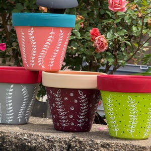 May include: A collection of four painted ceramic flower pots. Each pot features a different color scheme: coral with teal rim, gray with red rim, burgundy with cream rim, and green with red rim. White leaf patterns decorate the pots.